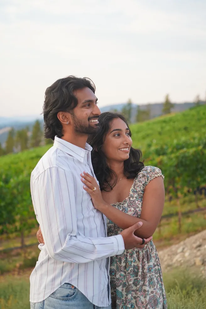 Neha and Shan embracing on a hillside vineyard, smiling into the distance