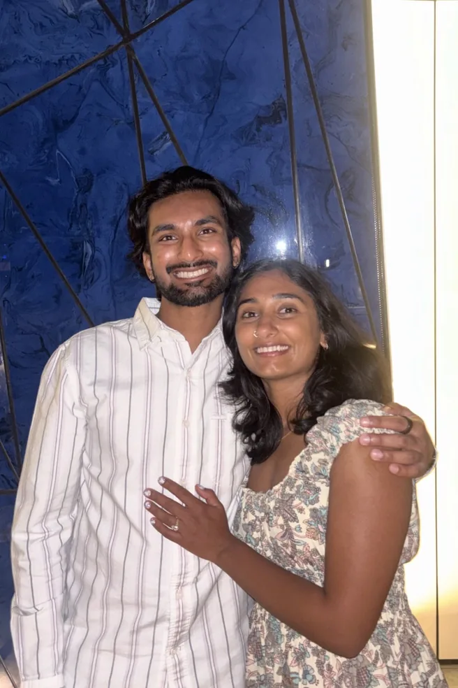 Neha and Shan smiling in front of a blue marble wall, Neha showing her ring